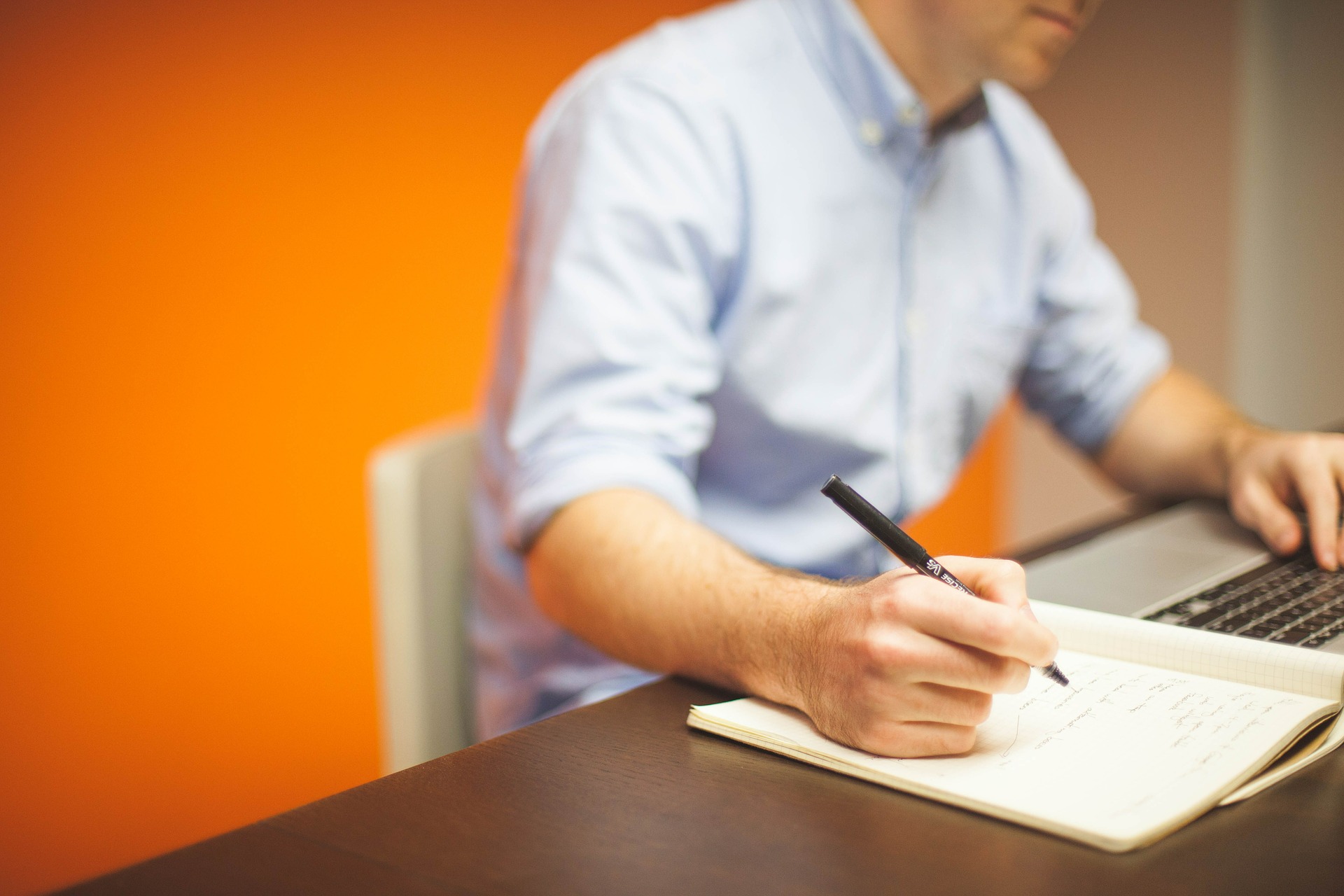 Man in blue button down writing in notebook against orange wall